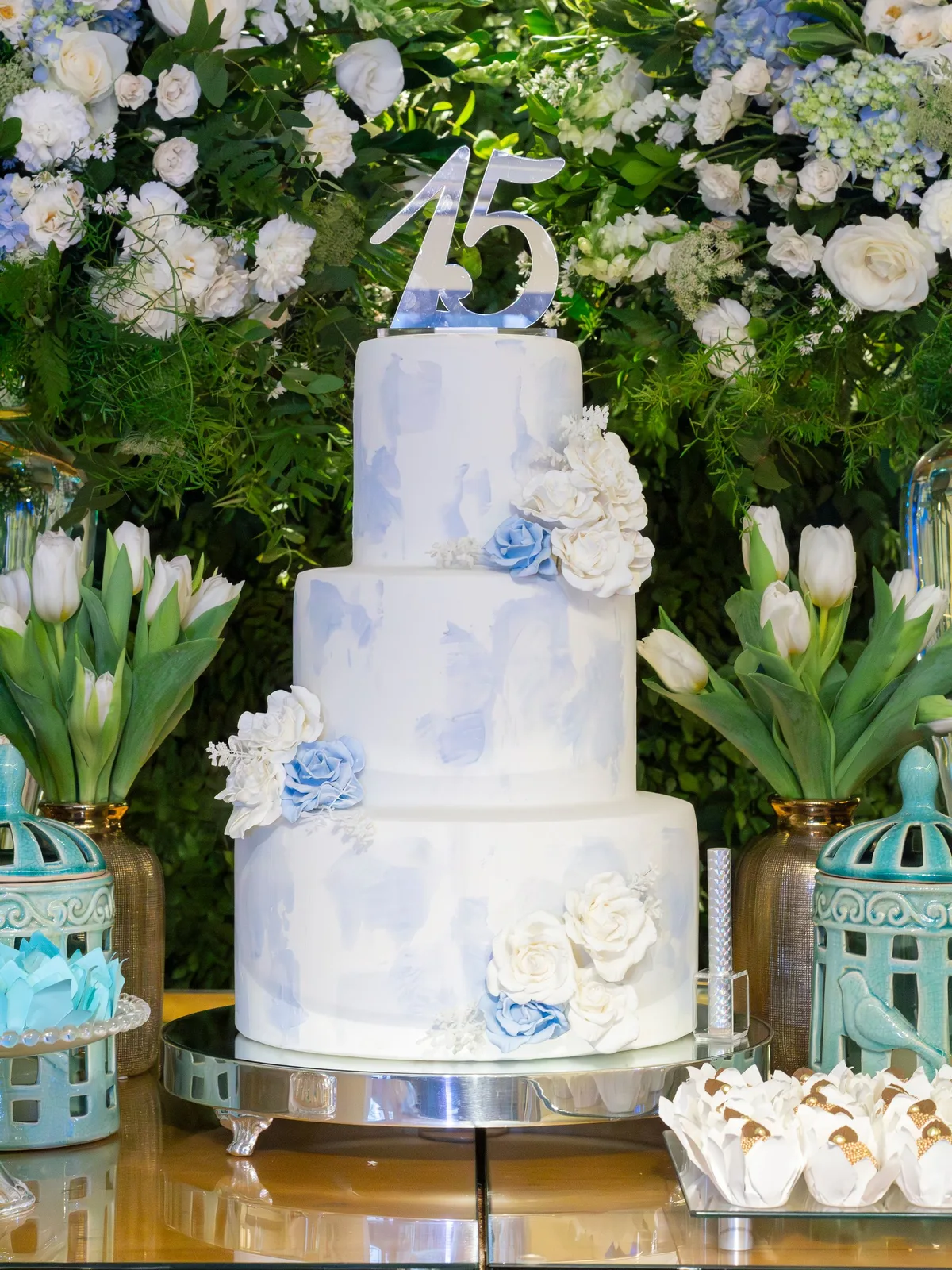 Pastel de quince años de tres pisos en azul y blanco, rodeado de arreglos de flores blancas